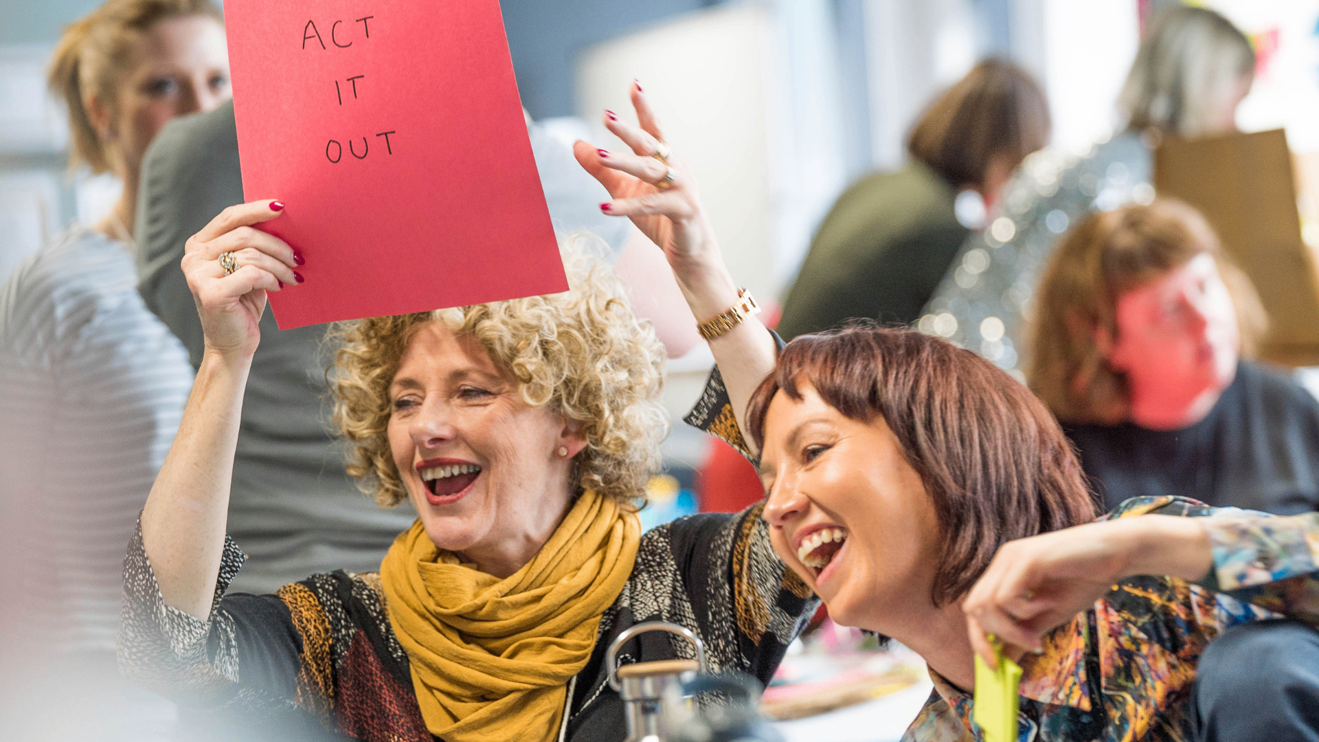 Workshop participants laughing while holding an 'Act It Out' prompt card